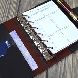 May include: Open leather planner with a pen and a credit card. The planner is black and brown with a silver binder. The pen is black and burgundy with gold accents. A blue Visa card is in the front pocket.