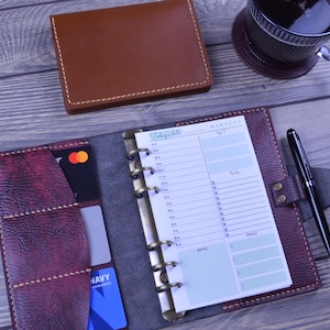 Op de afbeelding: Een open, bordeauxrode leren planner met een dagelijkse planningpagina, een zwarte pen en een bruine leren portemonnee. Een kopje koffie staat op een onderzetter op de achtergrond. Creditcards zijn zichtbaar.