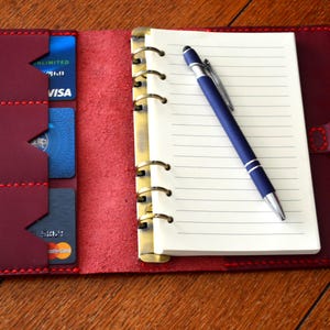 May include: A burgundy leather bound journal with a pen and card slots. The journal has a gold ring binder and red stitching.
