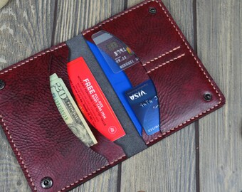 May include: A burgundy leather wallet with visible stitching and snap closures. The wallet is open, revealing a gray interior and pockets holding a twenty-dollar bill, credit cards, and a red promotional card. The wallet is on a wooden surface.