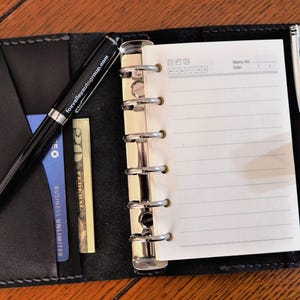 May include: A black leather bound planner with a pen and a credit card in the pocket. The planner has a silver ring binder and a pen holder.