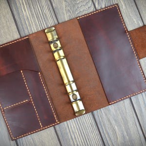 May include: Open, brown leather journal with a brass binder mechanism. The journal has a flap closure with two snap closures. The interior features pockets and is stitched with light-colored thread.