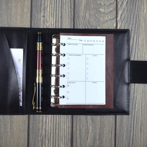 May include: Black leather planner with a gold button clasp, open to reveal a lined paper insert and a pen. The pen is black, gold, and burgundy. The planner is on a grey wooden surface.