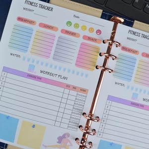 May include: A fitness tracker planner with a rose gold binder. The pages include sections for tracking meals, water intake, and workout plans. The planner has colorful boxes for breakfast, lunch, dinner, and snack, with a section for exercise type.