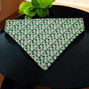 May include: A white triangular dog bandana with a green shamrock pattern. The bandana is decorated with a repeating pattern of green shamrocks and gold accents.