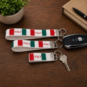 May include: Three fabric keychains featuring a repeating pattern of the Italian flag colors—green, white, and red—with the word ITALY printed in red. Each keychain has a silver metal clasp and a circular ring, with two holding keys.