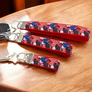 May include: Three red, white, and blue keychains with a repeating pattern of a man in a red cap saluting. The keychains are different sizes and have silver key rings.