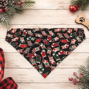 May include: A triangular pet bandana featuring a black background with a repeating pattern of Christmas stockings in various designs, including red, green, and white colors with stripes, snowflakes, and geometric shapes throughout the fabric.