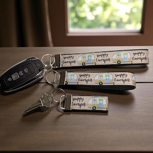 May include: Three keychains with the text "Happy Camper" and a camper illustration. One keychain is attached to a car key fob, another to a key, and the third is a wristlet. The keychains are beige, black, and white.