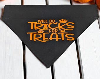 Puede incluir: Bandana triangular negra para perros con texto naranja "Will Do Tricks For Treats" y detalles de huellas de patas. La bandana está sobre una superficie de madera blanca, con pequeñas calabazas naranjas en el fondo. Un accesorio para mascotas con temática de Halloween.