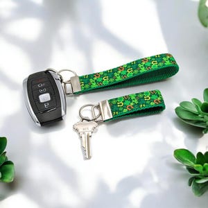 Op de afbeelding: Twee groene sleutelhangers met zilveren sleutelringen en een zilveren sleutel. De sleutelhangers zijn versierd met een patroon van groene klavers en gouden hoefijzers.