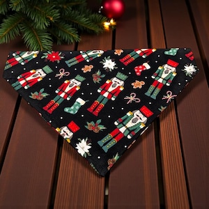 May include: Black dog bandana with a repeating pattern of nutcrackers in red and green uniforms, along with gingerbread men, stockings, and snowflakes. The bandana is triangular and lies on a wooden surface, with Christmas decorations in the background.