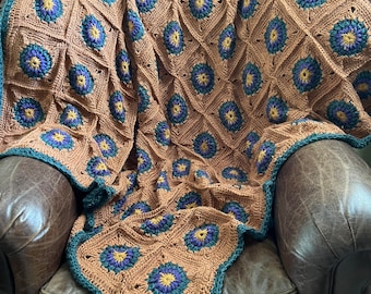 May include: A crocheted throw blanket with a brown background and a repeating pattern of green, purple, and yellow flowers. The blanket is draped over a brown leather armchair.
