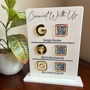 May include: A white acrylic sign with gold accents and the text "Connect With Us." It features gold icons for Google, Facebook, and Instagram, each paired with a QR code and the handle @dochefamilydental. A small plant is in the background.