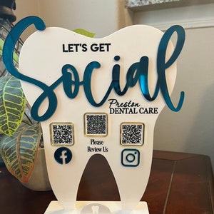 May include: A white, tooth-shaped sign with teal lettering that reads "Let's Get Social" and "Preston Dental Care." It features QR codes for Facebook, a review platform, and Instagram. The sign is designed to stand upright.