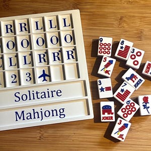 May include: A white Solitaire Mahjong game set with lettered and numbered tiles. The game board has slots for tiles and the words "Solitaire" and "Mahjong" printed on it. Scattered tiles feature various symbols and numbers.