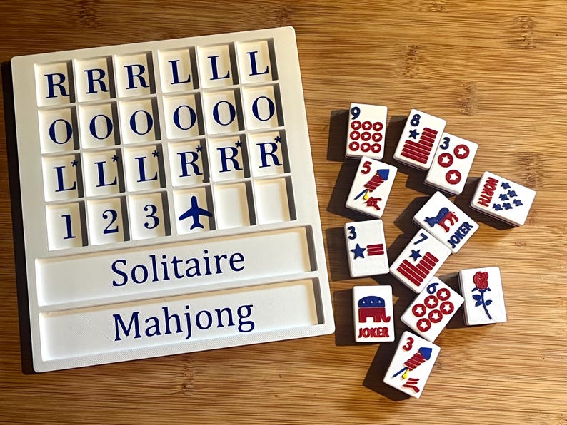 May include: A white Solitaire Mahjong game set with lettered and numbered tiles. The game board has slots for tiles and the words "Solitaire" and "Mahjong" printed on it. Scattered tiles feature various symbols and numbers.