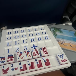 May include: A white board game with blue and red tiles. The tiles feature numbers, stars, and symbols like an airplane and a donkey. The game is set on a tray with lettered tiles. The word "JOKER" is visible on two tiles. The word "SOUTH" is also visible.