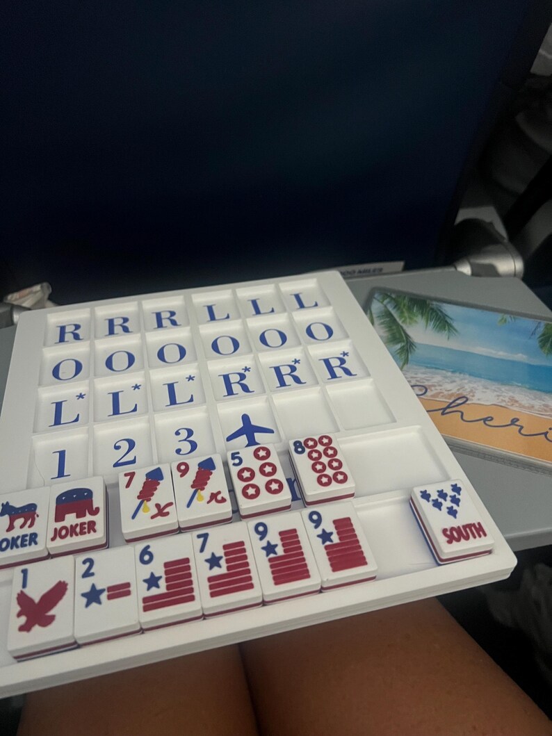 May include: A white board game with blue and red tiles. The tiles feature numbers, stars, and symbols like an airplane and a donkey. The game is set on a tray with lettered tiles. The word "JOKER" is visible on two tiles. The word "SOUTH" is also visible.