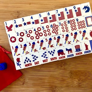May include: A set of white mahjong tiles featuring red and blue patriotic graphics including stars, stripes, fireworks, eagles, and political symbols. The tiles are arranged in a grid alongside two blue dice and a small red drawstring storage bag.