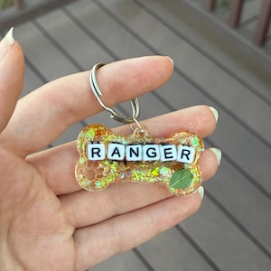 Può includere: Un portachiavi a forma di osso in resina traslucida con glitter dorati e scaglie iridescenti. Il portachiavi presenta il nome RANGER scritto con perline quadrate bianche con lettere nere e include una piccola foglia verde incastonata.