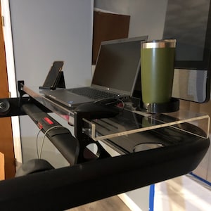 May include: A clear acrylic desk shelf attached to a black treadmill. A laptop, a smartphone, and a green travel mug are on the shelf.