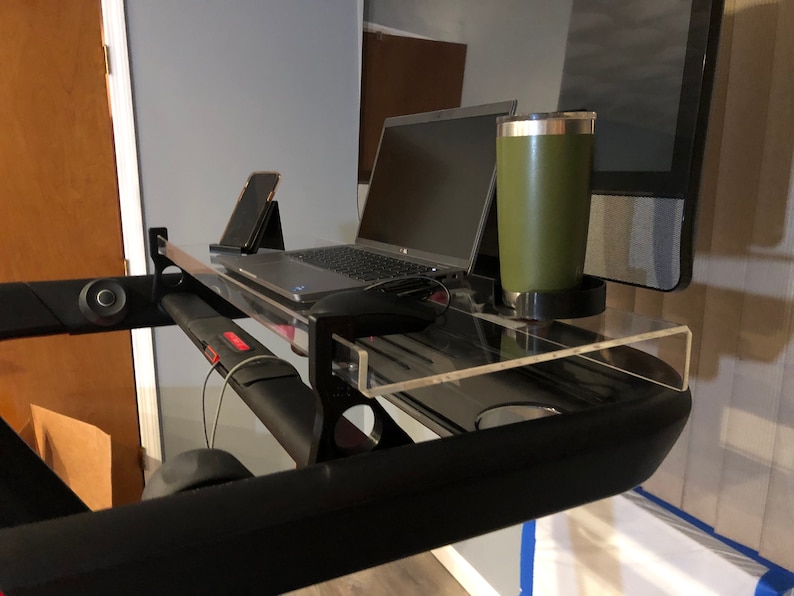 May include: A clear acrylic desk shelf attached to a black treadmill. A laptop, a smartphone, and a green travel mug are on the shelf.