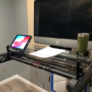 May include: A black treadmill with a clear acrylic desk attached. The desk has a tablet, a computer monitor, a piece of paper, and a green travel mug on it.