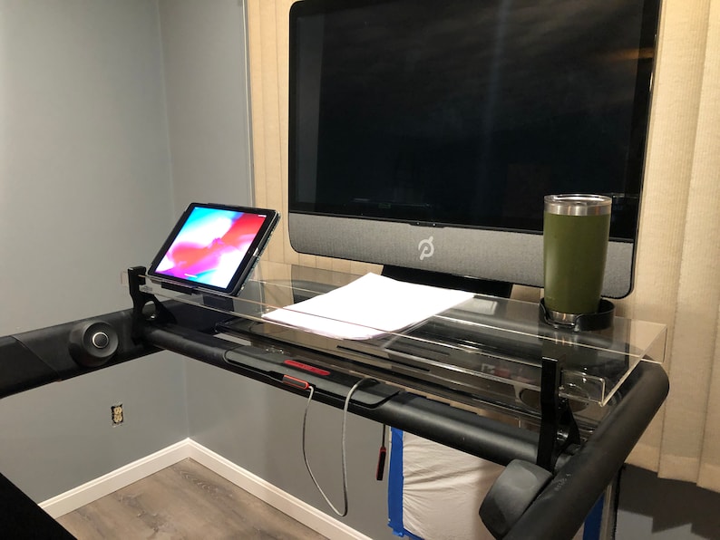 May include: A black treadmill with a clear acrylic desk attached. The desk has a tablet, a computer monitor, a piece of paper, and a green travel mug on it.
