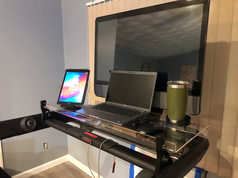 May include: A black treadmill with a clear acrylic desk attached. The desk has a laptop, tablet, and a green travel mug on it.