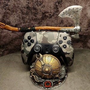 Op de afbeelding: Een PlayStation 5-controller met camouflagepatroon is gemonteerd op een standaard met een zilveren bijl en een gouden schild. Het schild heeft een gestileerd "Q"-symbool in het midden.