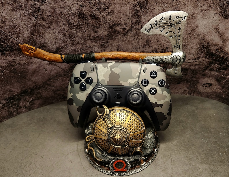 Op de afbeelding: Een PlayStation 5-controller met camouflagepatroon is gemonteerd op een standaard met een zilveren bijl en een gouden schild. Het schild heeft een gestileerd "Q"-symbool in het midden.