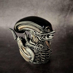 May include: Black and silver alien rubber duck with a detailed head and body. The duck has a black head with a silver mouth and teeth. The body is silver with black details.