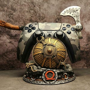 Op de afbeelding: Een PlayStation 5-controller met camouflagepatroon, een houten handvat, een zilveren bijl en een gouden schild, allemaal gemonteerd op een zwarte standaard met een rood "Q"-logo.