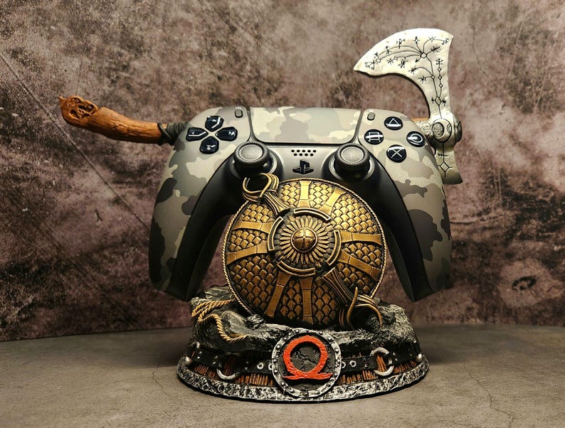 Op de afbeelding: Een PlayStation 5-controller met camouflagepatroon, een houten handvat, een zilveren bijl en een gouden schild, allemaal gemonteerd op een zwarte standaard met een rood "Q"-logo.