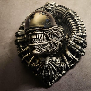 Puede incluir: Un busto de alienígena negro y plateado con una cara detallada y un diseño texturizado y orgánico. El busto está rodeado por un anillo de apéndices alienígenas.