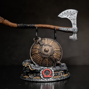May include: A display stand with a silver axe, a brown wooden staff, and a round, gold shield with a red and black symbol. The shield has a raised, circular design with a central point.