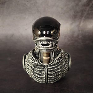 Puede incluir: Un busto de alien negro y plateado con una superficie detallada y texturizada. El alien tiene una cabeza negra con forma de casco y una boca grande abierta.