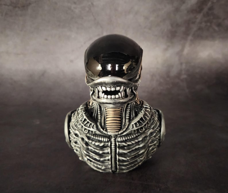 Puede incluir: Un busto de alien negro y plateado con una superficie detallada y texturizada. El alien tiene una cabeza negra con forma de casco y una boca grande abierta.