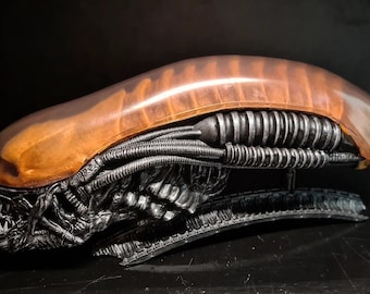 Puede incluir: Una escultura detallada de cabeza de alienígena, negra y plateada, con una cúpula naranja translúcida en la parte superior. La escultura está hecha de resina y es un objeto de colección popular para los fanáticos de la ciencia ficción.