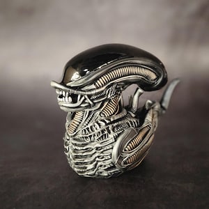 Puede incluir: Una figura de extraterrestre negra y plateada con un cuerpo segmentado detallado y una cabeza negra brillante. La figura est&aacute; en posici&oacute;n agachada con la cabeza inclinada hacia un lado.