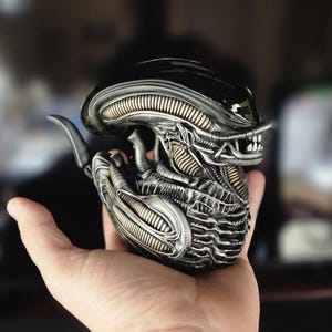 Puede incluir: Una figura de extraterrestre negra y plateada con un cuerpo segmentado detallado y una cabeza negra brillante. La figura est&aacute; en posici&oacute;n enroscada con la cabeza inclinada hacia un lado.