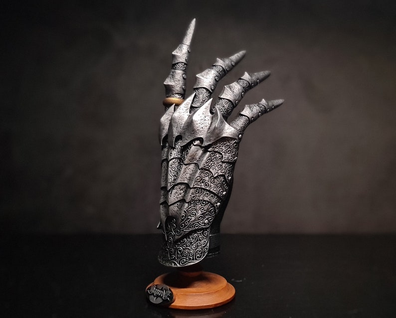 May include: A silver metal hand sculpture with intricate details and a ring on the index finger. The hand is posed with the fingers extended and the palm facing outward. The sculpture is mounted on a wooden base.