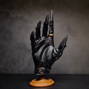 May include: A black leather glove with long, sharp claws and a gold ring on the index finger. The glove is mounted on a wooden stand.