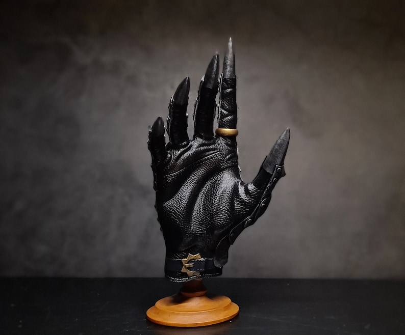 May include: A black leather glove with long, sharp claws and a gold ring on the index finger. The glove is mounted on a wooden stand.