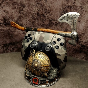 Op de afbeelding: Een camouflage-gekleurde videogamecontroller met een houten handvat en een zilveren bijl met ingewikkelde houtsnijwerk. De controller is gemonteerd op een basis met een gouden schildontwerp.