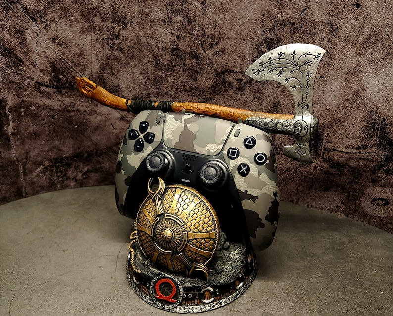 Op de afbeelding: Een camouflage-gekleurde videogamecontroller met een houten handvat en een zilveren bijl met ingewikkelde houtsnijwerk. De controller is gemonteerd op een basis met een gouden schildontwerp.