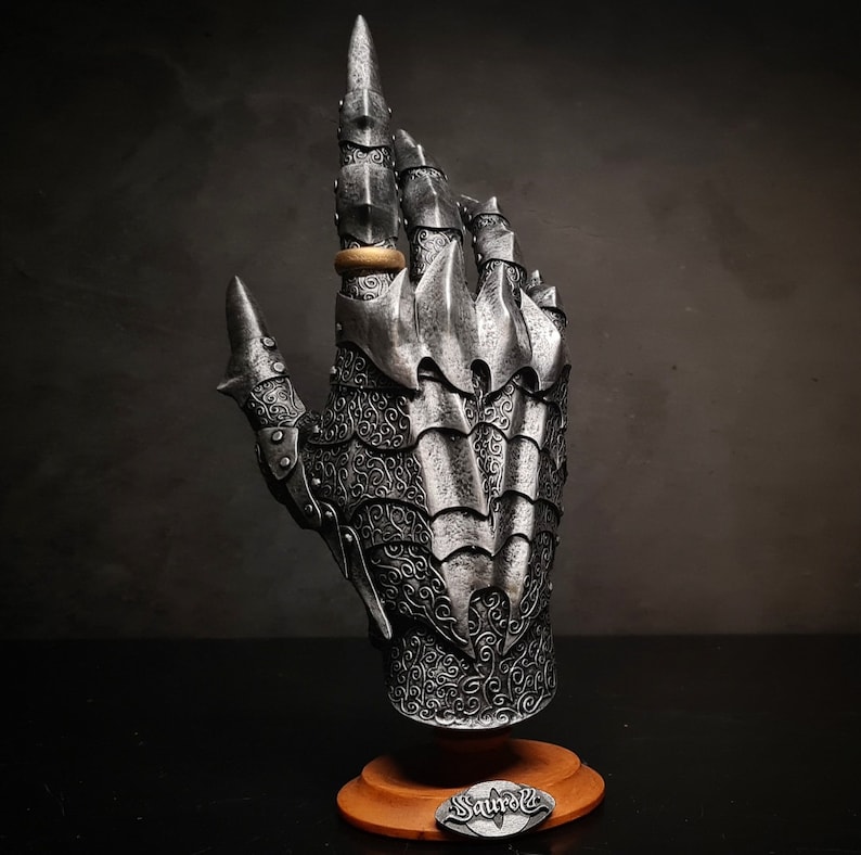 May include: A silver metal hand sculpture with intricate details, resembling a gauntlet or armor. The hand is wearing a gold ring and is mounted on a wooden base with the word "Saurol" inscribed on it.