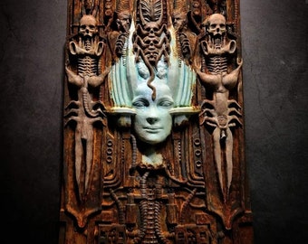 Puede incluir: Una escultura de pared de metal oxidado con un diseño detallado de un rostro humano rodeado de criaturas alienígenas. El rostro está pintado de color azul claro y la escultura tiene un aspecto texturizado y envejecido.