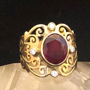 May include: A gold ring with a large, dark red gemstone surrounded by smaller, clear gemstones. The ring has a delicate, filigree design.